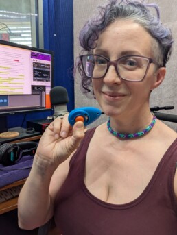 Arielle DeLisle, a white woman with short purple curly hair, sits in her recording studio, holding a blue and orange dog clicker device. Behind her is studio equipment, including a microphone, headphones and her monitor, displaying audio waveforms with narration and small areas of overmodulated audio from the dog clicker, indicating insertion points for the editor to work with.